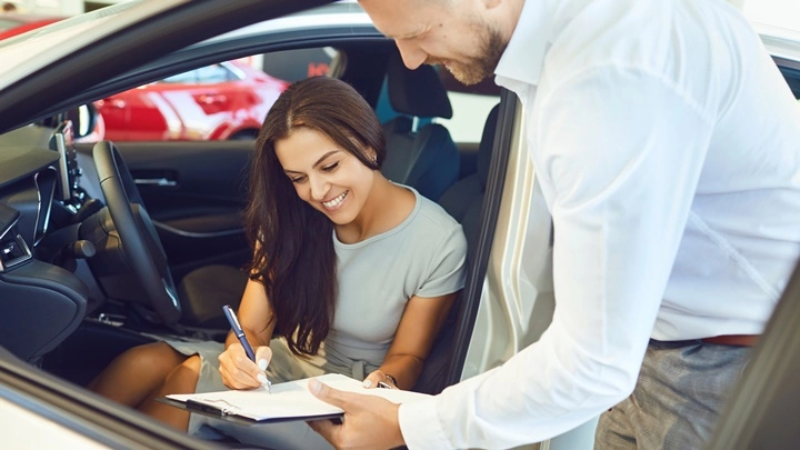 Eine Frau sitzt in einem Auto und unterschreibt einen Kaufvertrag, den ihr ein Autoverkäufer hinhält